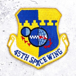 May include: An embroidered patch featuring a shield divided vertically into yellow and blue sections. It displays a globe graphic with a smaller moon, red flight symbols, and a white banner at the bottom with the text 45TH SPACE WING in blue.