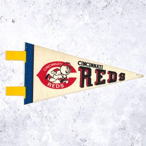 May include: A vintage Cincinnati Reds pennant in cream, red, black, blue, and yellow. The pennant features the team name and logo, including a running mascot. The pennant has a blue border and yellow tabs.