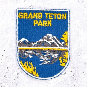Puede incluir: Parche bordado con el texto "GRAND TETON PARK" en amarillo sobre fondo azul. El parche presenta una cordillera reflejada en un lago, con detalles amarillos y blancos. El parche tiene forma de escudo.