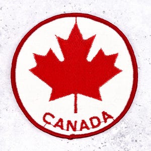 May include: Circular patch featuring a red maple leaf on a white background, encircled by a red border. The word "CANADA" is embroidered in red at the bottom. This patch is likely a souvenir or emblem.