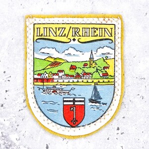 May include: A shield-shaped embroidered patch with a yellow border featuring a colourful illustration of the town of Linz am Rhein. The design includes a river with a paddle steamer and sailboat, a townscape with a church, and a red shield with a white key.