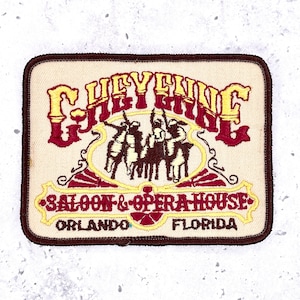 May include: A rectangular embroidered patch with a cream background and a dark brown border. It features the text CHEYENNE in yellow and red, an illustration of three figures on horseback, and the text SALOON & OPERA HOUSE ORLANDO FLORIDA.