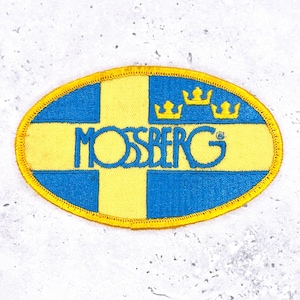 May include: An oval-shaped patch with a yellow border. The patch features a blue and yellow design with a cross, three crowns, and the word "MOSSBERG" in blue. The patch is likely an emblem or logo.