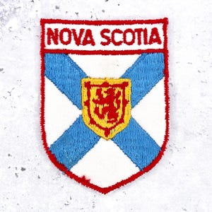 May include: A shield-shaped embroidered patch featuring the text NOVA SCOTIA in red letters on a white background. The design includes a blue saltire cross and a central yellow shield displaying a red lion, all bordered by a thick red stitched edge.