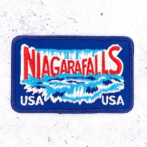 May include: A rectangular embroidered patch with a dark blue background. The text NIAGARAFALLS is stitched in red with a white outline above a stylized depiction of a waterfall in light blue and white. The text USA is stitched in white at the bottom.