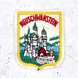 May include: Embroidered patch featuring Neuschwanstein Castle, with the castle in white, green, and red, set against a cream background. The patch has a yellow border and the castle name in red.