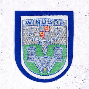 Könnte beinhalten: Blau-weißer Windsor-Aufnäher mit Schilddesign. Der Aufnäher zeigt oben das Wort "WINDSOR", einen Hirsch mit Geweih und zwei Burgtürme. Der Aufnäher ist mit einem blauen Stoff eingefasst.