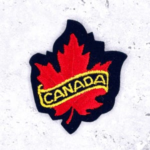 May include: Embroidered patch featuring a red maple leaf with the word "CANADA" in yellow lettering on a black background. The patch is in the shape of a maple leaf, a symbol of Canada.