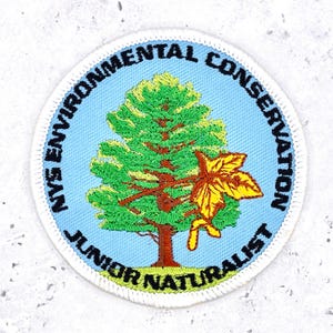 May include: A circular embroidered patch with a light blue background featuring a green pine tree and a yellow maple leaf. The text reads NYS ENVIRONMENTAL CONSERVATION JUNIOR NATURALIST in black lettering along the border of the patch.