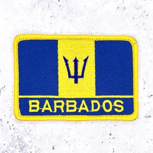May include: A rectangular embroidered patch featuring the Barbados flag design with two vertical blue bands flanking a central yellow band. A blue trident head is centered in the yellow section, and the word BARBADOS is stitched in yellow below.
