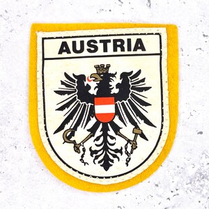 May include: A rectangular patch with a yellow border and the word "AUSTRIA" at the top. The patch features the Austrian coat of arms, including a black eagle with a red and white shield.