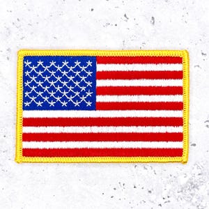 May include: Embroidered rectangular patch featuring the American flag. The design includes a blue field with white stars and red and white stripes. The patch is bordered with a yellow stitched edge.