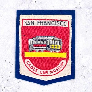 Op de afbeelding: Een rechthoekige geborduurde patch met een blauwe rand en een crèmekleurig midden. De patch heeft een rood schildontwerp met een zwart-gele kabelbaanillustratie, een gele horizontale streep en de tekst SAN FRANCISCO CABLE CAR MUSEUM.
