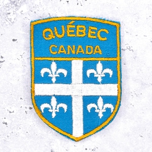 May include: A shield-shaped embroidered patch featuring a blue background with a yellow border. The text QUÉBEC CANADA is stitched in yellow at the top. Below, a white cross divides the blue field into four sections, each containing a white fleur-de-lis.