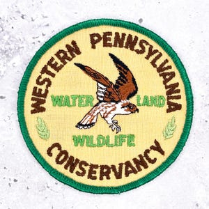 May include: A circular embroidered patch with a cream-colored background and a green border. It features a brown and white bird of prey in the center. The text reads WESTERN PENNSYLVANIA WATER LAND WILDLIFE CONSERVANCY in brown and green lettering.