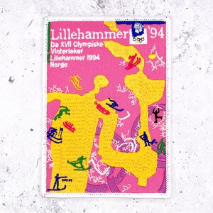 Könnte beinhalten: Ein rechteckiger gestickter Aufnäher mit rosa Hintergrund und einer großen gelben abstrakten Form. Er zeigt den Text Lillehammer '94, De XVII Olympiske Vinterleker, Lillehammer 1994, Norge sowie das Olympische Ringe-Logo mit einer blauen Flamme.