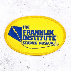 May include: An oval-shaped embroidered patch with a bright yellow background and a blue border. It features a blue kite graphic and the blue embroidered text THE FRANKLIN INSTITUTE SCIENCE MUSEUM, with a thin blue line connecting the kite to a key.