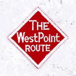 Pode incluir: Um patch bordado vermelho em forma de losango com costuras brancas. O patch apresenta o texto "THE WestPoint ROUTE" em branco, com letras estilizadas. O patch tem uma borda branca e está sobre um fundo cinza claro.