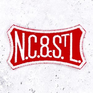 May include: A red embroidered patch with a white border and white stitched lettering that reads N.C.8.S.T.L. The patch has a curved, rectangular shape with flared corners and a dense, textured thread finish throughout the entire design.