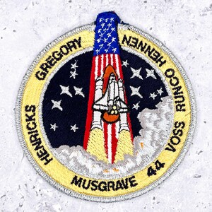 May include: Embroidered patch featuring a space shuttle launching into space, with the American flag at the top. The patch is circular, with the names of the astronauts around the edge and the number 44.