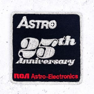 Op de afbeelding: Een zwarte vierkante patch met wit geborduurd tekst. De patch leest "ASTRO 25th Anniversary" en "RCA Astro-Electronics" in het rood. De patch heeft een zilveren rand.