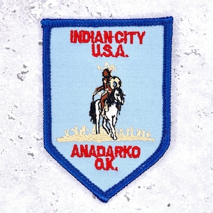 May include: A light blue embroidered patch with a shield shape. The patch features the text "INDIAN CITY U.S.A." in red, and "ANADARKO OK." in red. A Native American figure on horseback is in the center.