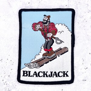 May include: A rectangular embroidered patch with a black border featuring a bearded character in a red plaid shirt and dark pants skiing on a snowy slope. The character holds a pole with a hook. The text BLACKJACK is printed in black at the bottom.