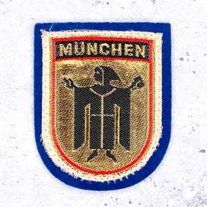 Op de afbeelding: Een blauwe, geborduurde patch met het woord "MUNCHEN" in rode letters bovenaan. De patch heeft een goud en zwart embleem met een gestileerde figuur. De patch is omrand met gouden en witte stiksels.