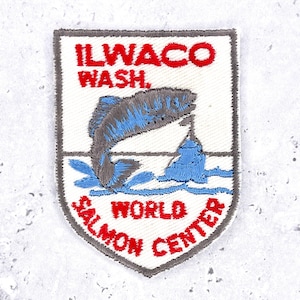May include: A shield-shaped embroidered patch with a white background and a gray border. It features red text reading ILWACO WASH. and WORLD SALMON CENTER. A gray and blue fish is depicted jumping over blue water waves in the center of the patch.