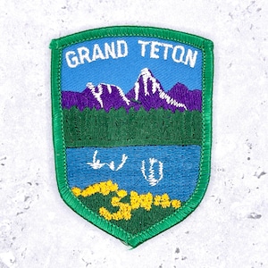 May include: A shield-shaped embroidered patch featuring the text GRAND TETON in white letters. The design depicts a blue sky, purple mountains with white snow-capped peaks, a dark green forest, a blue lake with white reflections, and yellow flowers.