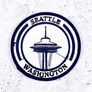 May include: A circular patch featuring the Space Needle in Seattle, Washington. The design is in navy blue on a white background, with the city and state name arched around the top and bottom edges in navy blue lettering. The patch has a navy blue border.