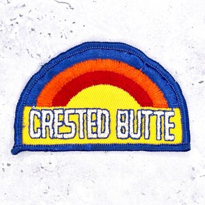 May include: A semi-circular embroidered patch featuring a vibrant rainbow design with concentric bands of orange and red over a bright yellow base. The text CRESTED BUTTE is stitched in bold white capital letters across the yellow section.