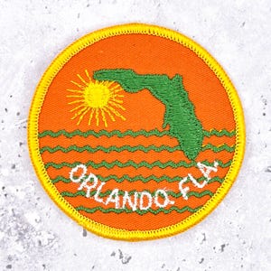 May include: Embroidered patch featuring a stylized depiction of Florida with a sun and water waves. The patch is orange with green and yellow accents. The text "ORLANDO, FLA." is embroidered in white.