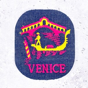 May include: A denim patch with a yellow gondola and gondolier, a pink bridge, and a striped pole. The word "VENICE" is in pink at the bottom. The patch has a dark blue background and an oval shape.