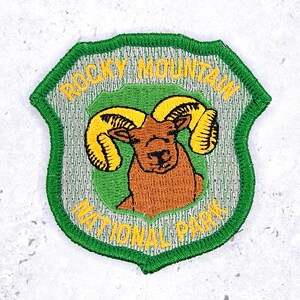 May include: Embroidered patch depicting a brown ram with yellow horns, within a green and yellow shield. The text "ROCKY MOUNTAIN NATIONAL PARK" is arched around the top and bottom of the design.