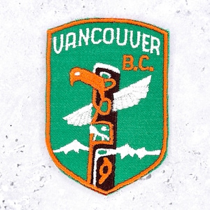 May include: A shield-shaped embroidered patch with a green background and orange border. It features the white text VANCOUVER B.C. above a brown totem pole graphic with an orange eagle head, white wings, and white mountain silhouettes.
