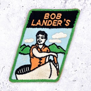 Könnte beinhalten: Gestickter Aufnäher mit dem Text "BOB LANDER'S" in Orange und Weiß auf schwarzem Hintergrund. Der Aufnäher zeigt einen Mann in einem orangefarbenen Hemd in einem Boot, mit blauem Himmel, Wolken und grünen Bergen.