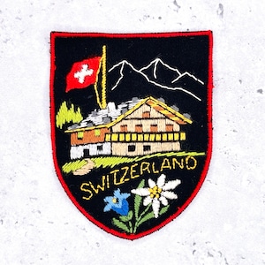 May include: Embroidered patch depicting a Swiss chalet, mountains, and the Swiss flag. The patch has a black background with red trim and the word "SWITZERLAND" in yellow.
