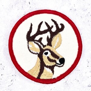 May include: A circular embroidered patch featuring a deer head with brown antlers and tan fur against a white background, enclosed by a thick red stitched border. The design shows the deer in profile, highlighting the dark brown facial markings.