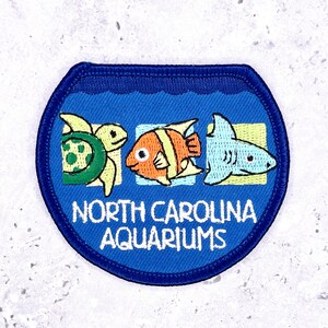 Könnte beinhalten: Blauer Aufnäher mit einer Schildkröte, einem Clownfisch und einem Hai. Der Aufnäher trägt den weißen Schriftzug "NORTH CAROLINA AQUARIUMS". Der Aufnäher hat eine runde Form mit blauem Rand.