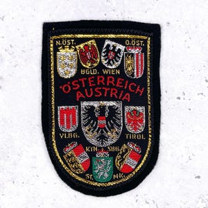 May include: A black embroidered patch with gold trim and the words "ÖSTERREICH AUSTRIA" in red. The patch features several colorful crests and the names of Austrian regions.
