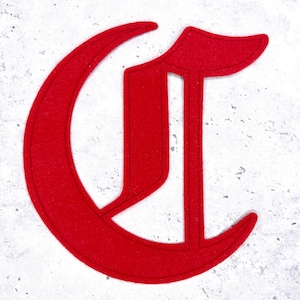 May include: A bright red felt letter "C" in a gothic font style. The letter is centered against a speckled white background. The edges of the letter are cleanly cut, and the overall design is simple and bold.