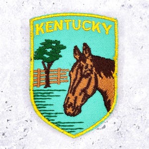 Könnte beinhalten: Gestickter Aufnäher mit dem Wort "Kentucky" in Gelb auf türkisfarbenem Hintergrund. Der Aufnäher zeigt einen Pferdekopf, einen Baum und einen Zaun. Der Aufnäher hat einen gelben Rand.