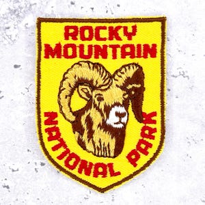 Op de afbeelding: Een schildvormige geborduurde patch met een felgele achtergrond en een donkerbruine rand. Het bevat een centrale illustratie van een dikhoornschaapskop in bruin en beige, met de rode tekst ROCKY MOUNTAIN NATIONAL PARK eromheen.
