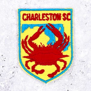May include: Embroidered patch featuring a red crab over a blue and yellow background. The text "CHARLESTON SC" is displayed in red at the top. The patch has a yellow border and is likely for apparel or accessories.