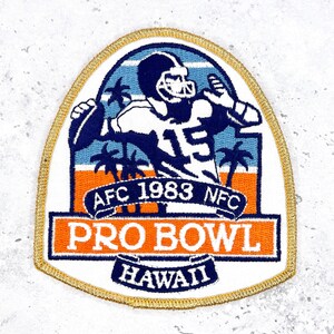 May include: Embroidered patch featuring a football player throwing a ball, set against a blue and orange tropical scene with palm trees. The patch reads "AFC 1983 NFC PRO BOWL HAWAII" in white and blue lettering, with a gold border.