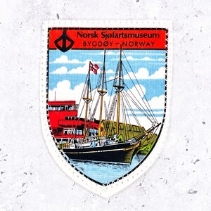 May include: A shield-shaped embroidered patch featuring a black sailing ship on blue water. The top red banner displays the text Norsk Sjøfartsmuseum BYGDØY - NORWAY. A red building stands in the background under a blue sky with white clouds.