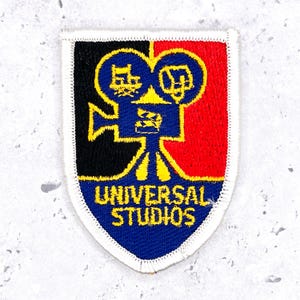 May include: A shield-shaped embroidered patch featuring a blue film camera icon with yellow outlines, set against a split black and red background. The text UNIVERSAL STUDIOS is embroidered in yellow at the bottom, all bordered by white stitching.