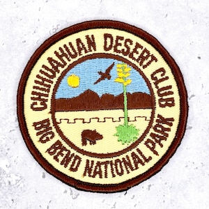 May include: A circular embroidered patch with a brown border and cream background. The text reads CHIHUAHUAN DESERT CLUB BIG BEND NATIONAL PARK. The center depicts a blue sky, brown mountains, a yellow sun, a bird, a cactus, and a brown bear.