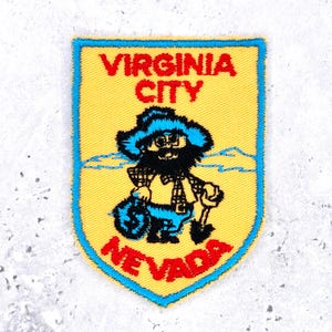May include: A shield-shaped embroidered patch with a yellow background and a blue border. It features the red text VIRGINIA CITY and NEVADA. A central graphic depicts a bearded prospector in a blue hat holding a shovel and a money bag.